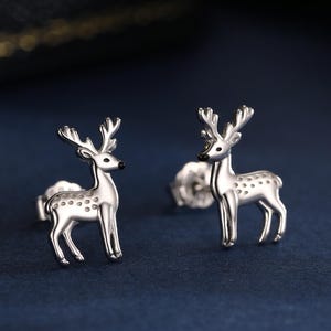 May include: A pair of silver deer stud earrings. Each earring has a detailed deer design with antlers and dotted markings. The earrings are presented on a dark blue surface, with a blurred background.