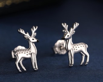 May include: A pair of silver deer stud earrings. Each earring has a detailed deer design with antlers and dotted markings. The earrings are presented on a dark blue surface, with a blurred background.