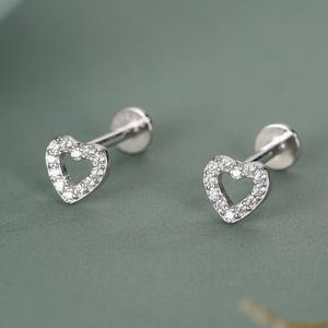 Puede incluir: Un par de pendientes de botón plateados, cada uno con un diseño en forma de corazón. El corazón está delineado con pequeñas piedras claras y brillantes. Los pendientes se muestran sobre una superficie verde apagada.