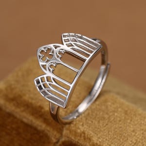 May include: A silver-toned metal ring featuring an openwork design that mimics the architectural structure of a gothic cathedral window, including pointed arches and a quatrefoil cutout detail at the top centre of the band.