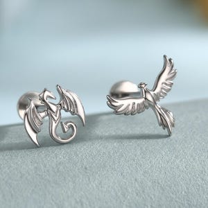 May include: A pair of silver-colored stud earrings. One earring features a dragon design, and the other depicts a phoenix in flight. The earrings are displayed on a gray surface with a soft focus background.