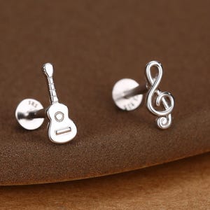 Könnte beinhalten: Ein Paar silberne Ohrstecker. Ein Ohrring ist wie eine kleine Gitarre geformt, der andere ist ein Violinschlüssel. Die Ohrringe befinden sich auf einer braunen Oberfläche.