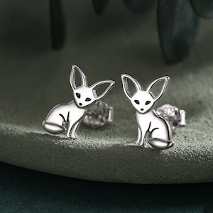 May include: A pair of silver-colored stud earrings featuring a stylized fox design. The earrings depict a sitting fox with large ears and a small, round body. The earrings are displayed on a textured, green surface.