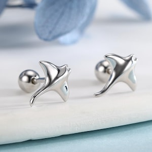 Op de afbeelding: Een paar zilverkleurige oorstekers. Elke oorbel heeft een gestileerd manta rog ontwerp met een glad, reflecterend oppervlak. De oorbellen zijn geplaatst tegen een witte en blauwe achtergrond, met blauwe bloemaccenten.