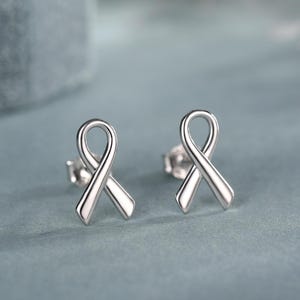 Könnte beinhalten: Ein Paar silberne Ohrstecker in Form von Schleifen. Die Ohrringe werden auf einer blau-grauen Oberfläche mit unscharfem Hintergrund präsentiert. Die Ohrringe sind ein Symbol für Bewusstsein und Unterstützung.