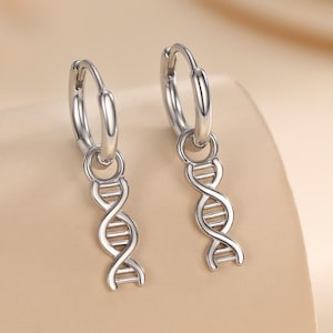 Könnte beinhalten: Ein Paar silberne Creolen mit einem DNA-Helix-Anhänger. Die Ohrringe bestehen aus glänzendem Metall und hängen an einem kreisförmigen Reifen. Der DNA-Helix-Anhänger ist eine detaillierte Darstellung der Doppelhelix-Struktur.