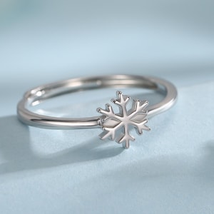 Puede incluir: Un anillo plateado con un diseño de copo de nieve. El anillo está sobre un fondo azul claro. El copo de nieve es el punto focal, con detalles intrincados. El anillo parece ser ajustable.