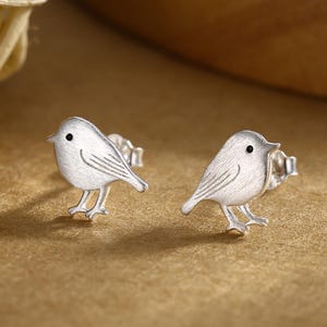 Op de afbeelding: Een paar zilveren oorstekers met vogelmotief. Elke oorbel heeft een klein, gedetailleerd vogelontwerp met een getextureerd lichaam, kleine pootjes en een klein zwart stipje voor het oog. Op een neutrale achtergrond.