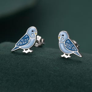 以下が含まれることがあります： 青いセキセイインコのモチーフの、銀色のスタッドピアス。ピアスは、明るい青色のボディに濃い青色の羽のディテールと黄色のくちばしが特徴です。ピアスは濃い緑色の背景に配置されています。