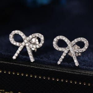 May include: A pair of silver bow-shaped stud earrings, each set with small, clear crystals. The earrings are presented on a dark blue velvet surface, with a blurred blue backdrop.