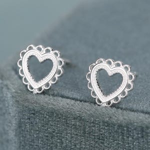May include: A pair of silver stud earrings, each featuring an open heart shape outlined by a delicate scalloped edge with small circular cutouts. The earrings have a polished metallic finish and are designed as simple, symmetrical heart-shaped studs.