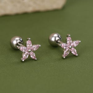 May include: A pair of silver-toned stud earrings, each featuring a pink, five-petal flower design. The earrings are set against a green background, with a textured, light-colored stone in the background.