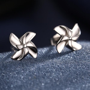 May include: A pair of silver-colored stud earrings. Each earring features a pinwheel design with curved blades. The earrings are displayed on a blue velvet surface, with a blurred background. The earrings are likely made of metal.