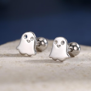 Puede incluir: Un par de pendientes de botón plateados con forma de fantasma. Cada pendiente presenta un pequeño fantasma sonriente con un cuerpo redondeado y un poste de bola. Los pendientes se muestran sobre una superficie texturizada blanquecina.