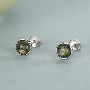 Puede incluir: Un par de pendientes de botón con engaste plateado y una piedra redonda de color verde oliva. Los pendientes se muestran sobre una superficie azul claro, con un fondo borroso. Diseño sencillo y elegante.