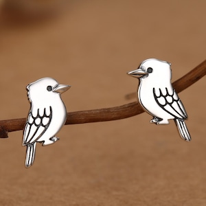 Könnte beinhalten: Ein Paar Ohrstecker in Form kleiner Vögel. Jeder Ohrstecker hat eine polierte Silberoberfläche mit eingravierten Linien, die die Flügel- und Schwanzfedern definieren, sowie einen kleinen schwarzen Punkt als Auge des Vogels.