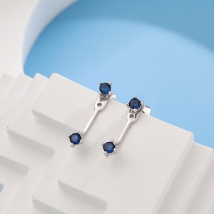 Könnte beinhalten: Ein Paar silberne Ohrringe mit einem kleinen blauen Edelstein an jedem Ende. Die Ohrringe sind so konzipiert, dass sie mit dem Edelstein nach vorne getragen werden, und der silberne Stift ist hinter dem Edelstein sichtbar.