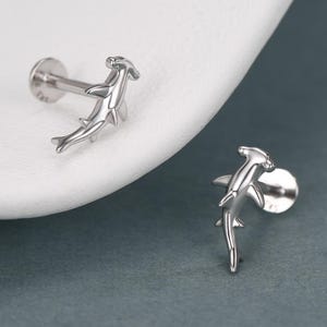 May include: A pair of silver-colored hammerhead shark stud earrings. The earrings are detailed with the shape of a hammerhead shark. The earrings are displayed on a white surface against a gray background.