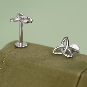 Könnte beinhalten: Zwei silberfarbene Ohrstecker mit einem keltischen Knoten-Design. Einer ist im Pfostenstil, der andere ein flacher Stecker. Die Ohrringe werden auf einer grünen Oberfläche vor einem weichen grünen Hintergrund präsentiert.