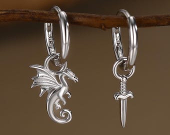 May include: A pair of silver hoop earrings. One earring features a dragon charm, the other a sword charm. The earrings are displayed on a brown branch against a blurred background.