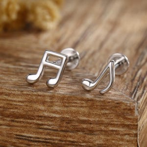 Peut inclure: Une paire de boucles d'oreilles argentées en forme de notes de musique. Les boucles d'oreilles sont présentées sur une surface en bois, avec un arrière-plan flou de fleurs séchées. Petites et délicates, elles sont parfaites pour les mélomanes.