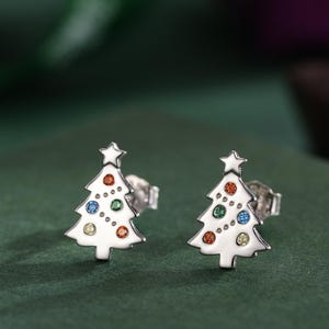 Könnte beinhalten: Ein Paar silberfarbene Ohrstecker in Form eines Weihnachtsbaums. Jeder Ohrring hat einen weißen Emaille-Baum mit bunten Punktakzenten und einem Stern. Die Ohrringe werden auf einer dunkelgrünen Oberfläche präsentiert.
