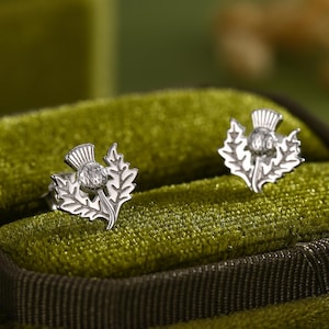 Op de afbeelding: Een paar zilverkleurige distel oorknopjes, gepresenteerd in een groene fluwelen sieradendoos. De oorbellen tonen het ontwerp van de Schotse nationale bloem met een centrale steen. De oorbellen liggen op een groene fluwelen ondergrond.