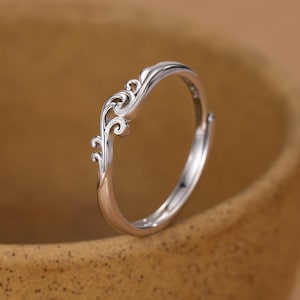 May include: A silver-coloured ring featuring a delicate, openwork design on the top band that resembles stylised, swirling wave patterns. The metal has a polished, reflective finish, and the band is thin, creating a lightweight appearance.