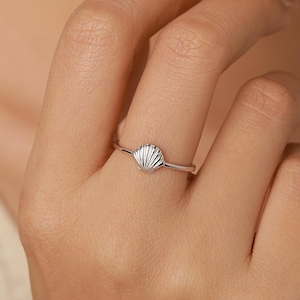 May include: A thin silver-toned ring featuring a small, textured seashell charm at the center. The band is smooth and metallic, showcasing a delicate design that sits horizontally across the finger with fine ridges etched into the shell surface.