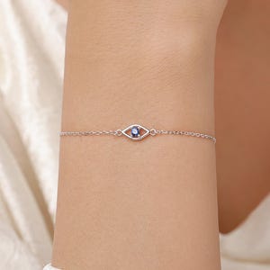 Op de afbeelding: Een delicate zilverkleurige kettingarmband met een centraal oogvormig bedeltje. Het bedeltje is omlijnd met zilverkleurig metaal en bevat een kleine, ronde, donkerblauwe edelsteen in het midden voor een verfijnde en elegante uitstraling.