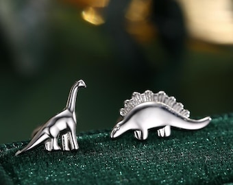 May include: A pair of silver dinosaur earrings. One earring features a long-necked dinosaur, while the other earring features a spiky dinosaur.