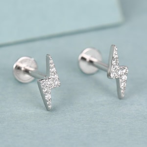 May include: A pair of silver stud earrings in the shape of lightning bolts. Each earring has a small, round back and is decorated with tiny, sparkling stones. The earrings are presented on a light blue surface.