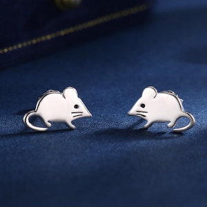 Op de afbeelding: Een paar zilverkleurige muisvormige oorstekers. Elke oorbel heeft een kleine, gestileerde muis met een gebogen staart en eenvoudige gezichtsdetails. De oorbellen worden gepresenteerd op een donkerblauwe ondergrond, met een fluwelen sieradendoos op de achtergrond.