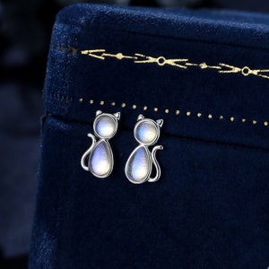 May include: A pair of silver cat-shaped earrings with iridescent moonstone insets. The earrings are displayed in a blue velvet jewellery box.