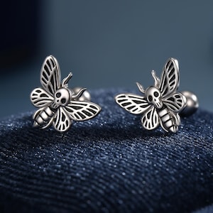 May include: A pair of silver stud earrings, each shaped like a moth with a skull motif. The moth's wings have detailed patterns. The earrings are presented on a dark blue, textured surface. The earrings are a unique accessory.