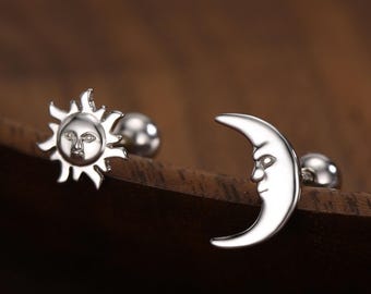 May include: Silver-colored mismatched earrings featuring a sun and a crescent moon design. The sun has a face in the center with rays extending outward. The moon has a face on the curved side. The earrings are on a wooden surface.