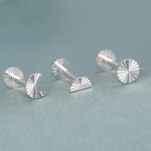 May include: Three silver-toned flat-back stud earrings featuring textured, sunburst-patterned designs. The set includes a crescent moon shape, a semi-circle shape, and a full circular shape, each with a straight post and a flat circular backing.