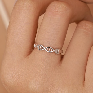 May include: A silver-coloured ring featuring a continuous, open-work design that mimics the double helix structure of a DNA strand. The band is thin and metallic, with repeating horizontal rungs connecting two intertwined, curved lines.
