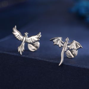 Könnte beinhalten: Ein Paar silberfarbene Ohrstecker. Ein Ohrring zeigt einen Phönix mit ausgebreiteten Flügeln, der andere einen Drachen mit ausgebreiteten Flügeln. Die Ohrringe werden auf einer dunkelblauen Oberfläche präsentiert.