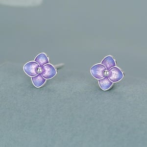 以下が含まれることがあります： 4枚の花びらを持つ花のデザインのスタッドピアス一対。各ピアスは銀色の金属製の縁取りが施され、中央には紫と青のグラデーションのエナメルが塗られており、中心には小さな銀色のビーズがあしらわれています。