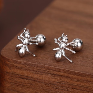 Peut inclure: Deux boucles d'oreilles clous argentées en forme de fourmi. Chaque boucle d'oreille présente un design de fourmi détaillé avec six pattes et un dos rond. Les boucles d'oreilles sont présentées sur une surface en bois foncé.