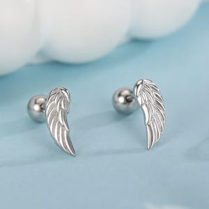 May include: A pair of silver-colored stud earrings, each featuring a detailed wing design. The earrings are set against a light blue background with white orbs and a logo in the bottom right corner.