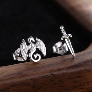 Op de afbeelding: Een paar zilveren oorbel studs, een met een drakenontwerp en de andere met een zwaardontwerp.