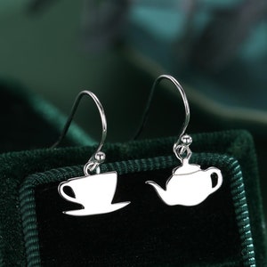 Pode incluir: Um par de brincos de prata com um design de bule de chá e xícara de chá. Os brincos são exibidos em um fundo de veludo verde.