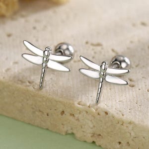 May include: A pair of silver dragonfly stud earrings with white enamel wings. The earrings are displayed on a textured, light-colored surface with a soft green background. The earrings are small and delicate, with detailed dragonfly designs.