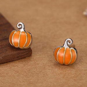 May include: A pair of stud earrings shaped like small pumpkins. Each earring features a bright orange enamel finish with polished silver-toned metal outlines and a small, curled stem at the top, creating a detailed miniature vegetable design.