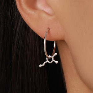 May include: A silver hoop earring featuring a small, dangling charm shaped like a chemical molecular structure. The earring is thin and circular, with the metallic charm hanging from the bottom of the hoop, displaying a distinct geometric pattern.