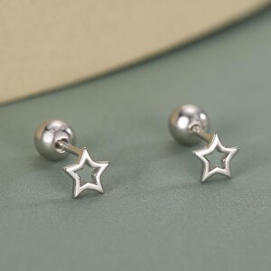 Könnte beinhalten: Ein Paar silberfarbene Stern-Ohrstecker. Jeder Ohrring hat ein kleines, offenes Sterndesign, das an einem Stecker mit rundem Verschluss befestigt ist. Die Ohrringe werden auf einer gedämpften grünen Oberfläche präsentiert.