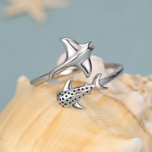 May include: A silver-toned open ring featuring a manta ray design on one end and a whale shark with black dotted detailing on the other end. The band is adjustable and crafted from polished metal, showcasing marine life motifs in a wrap-around style.