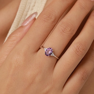 May include: A silver ring featuring a central oval-cut purple amethyst gemstone flanked by two small, clear, round-cut accent stones on a thin, polished metal band. The ring is worn on a finger, showcasing the prong setting of the purple stone.